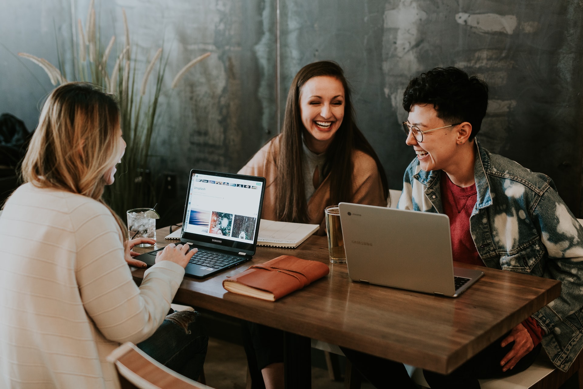 Three young people sit and laugh with one another with laptops, notebooks, and drinks on their table.