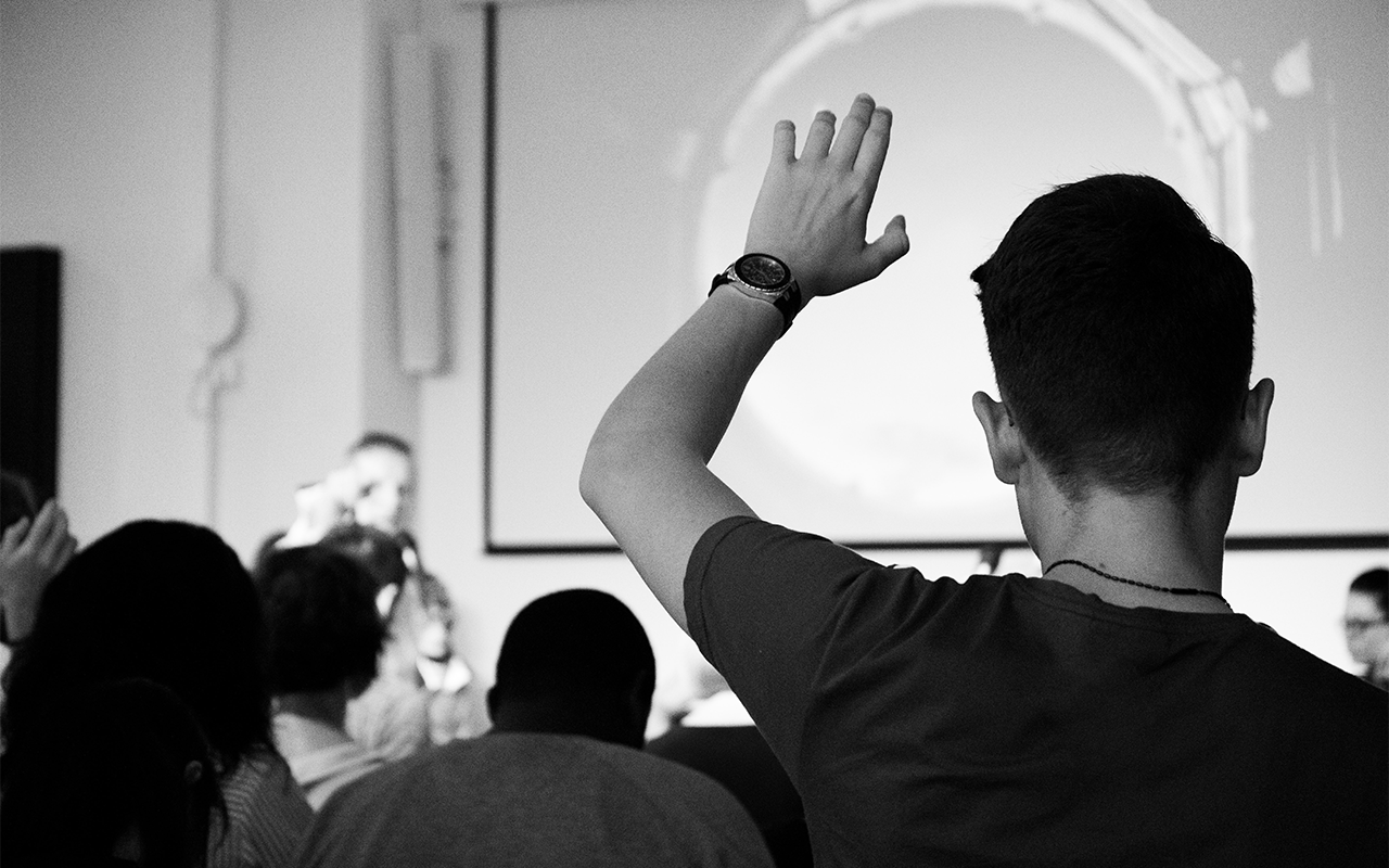 A black and white image looking to the front of the class. A learner raises their hand in the foreground.