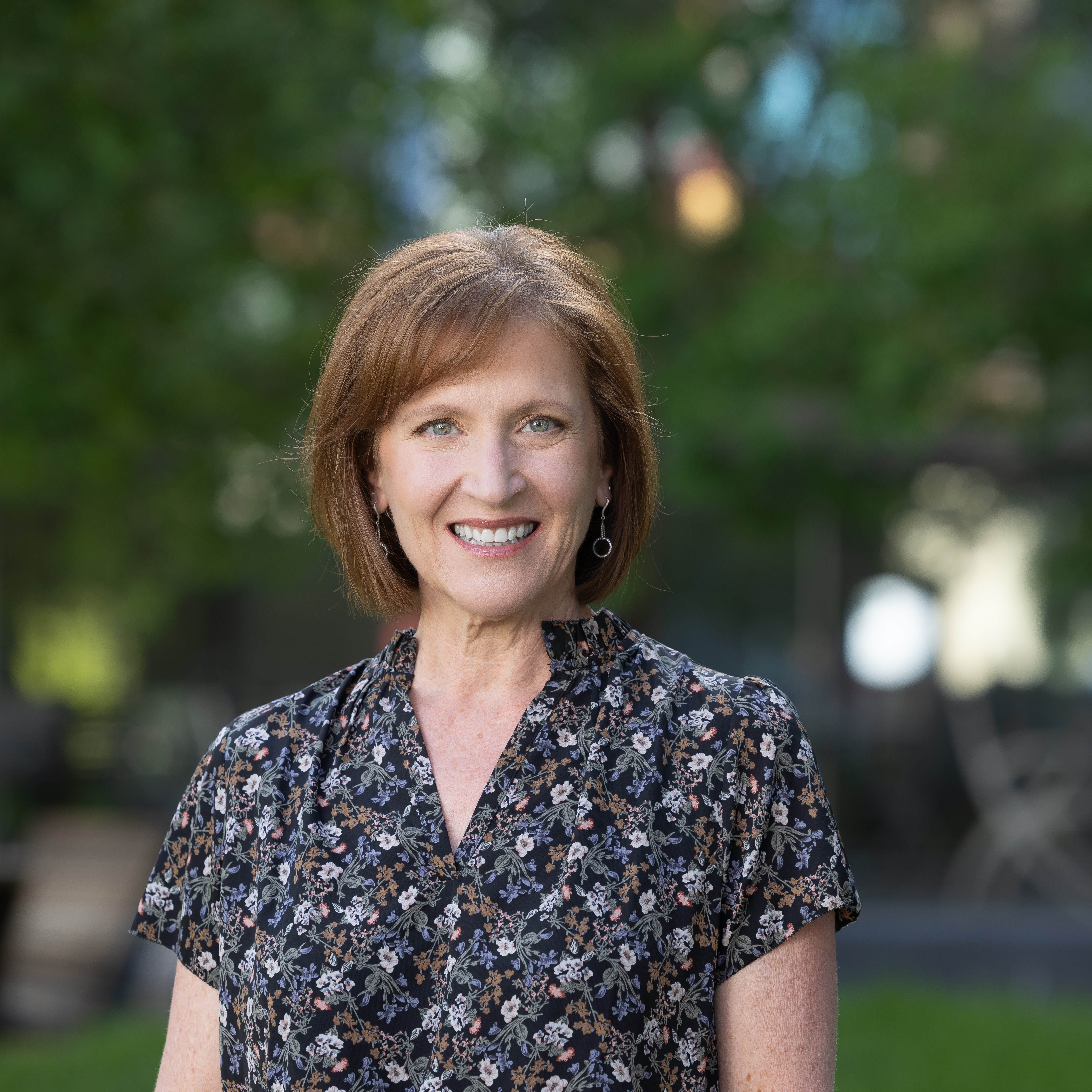 Professional photo of Elizabeth in front of trees. Elizabeth is a white woman with straight, short red hair. Elizabeth is wearing a black floral shirt and silver earrings.