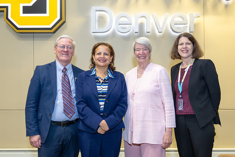 Four individuals in business attire smile in front of a lit sign reading 