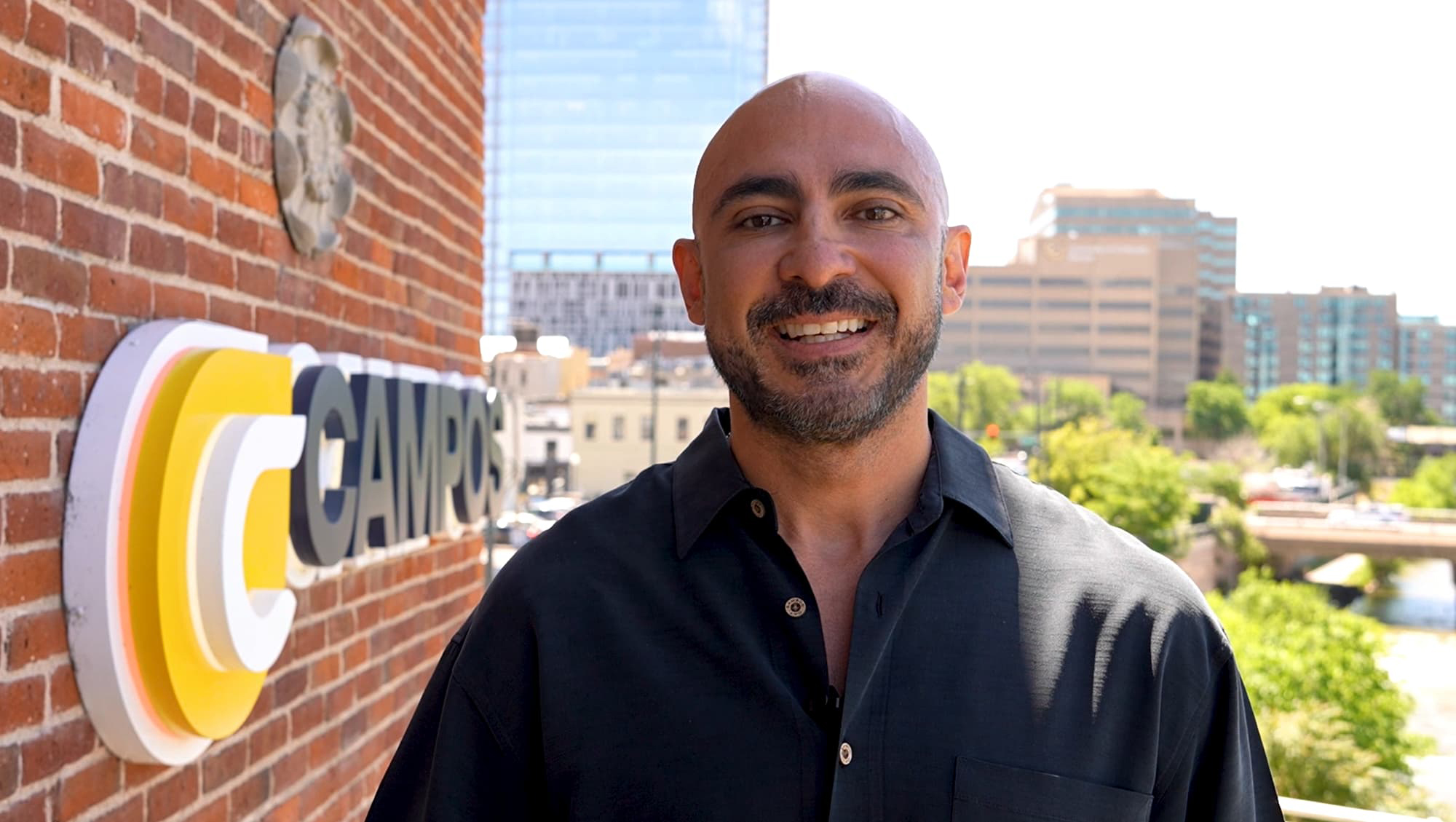 Marco Campos wearing a black polo and smiling with his teeth stands outside against a brick wall with the Campos branding.