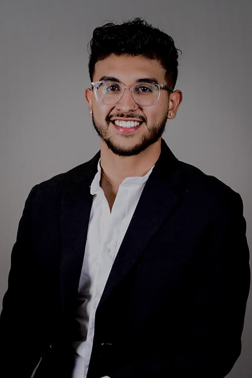 Oscar Fraire in a white button up and dark jacket, with clear-rimmed glasses, pierced ears and nose, smiling with his teeth. 