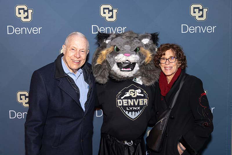 Two people dressed in professional attire smile with Milo the Lynx and a CU Denver backdrop behind them.