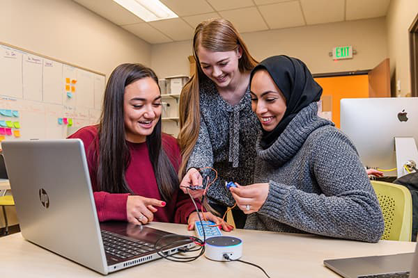 Three engineering students work on a project with wires and a lap top on the table, all smiling and interacting. 