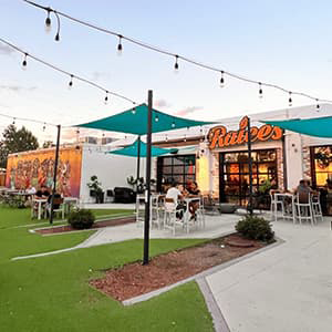 Outdoor patio of Racies Brewery Company with a bight mural, green AstroTurf, teal canopies, strung bistro lights, and people seated at tables.