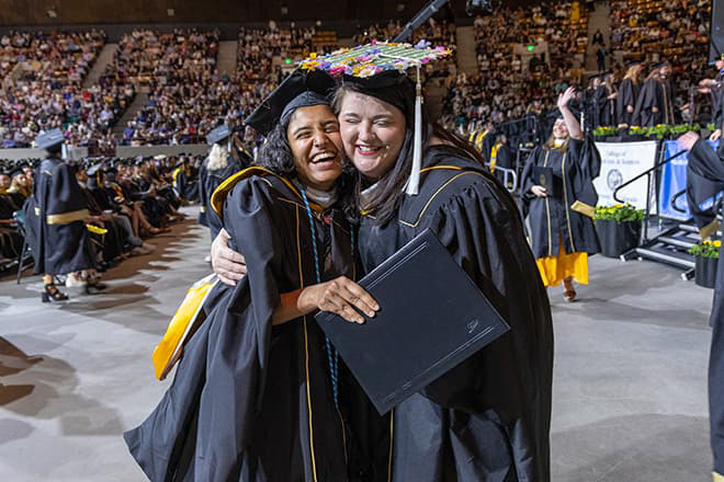 Two graduates in commencement regalia hugging each other with big, open-mouthed smiles.
