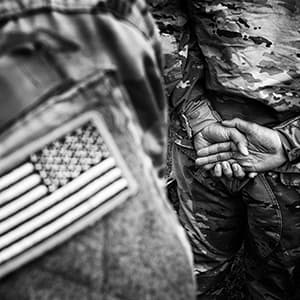 Black and white close up of a military uniform's flag and a the back of someone standing in the background, hands folded behind them.
