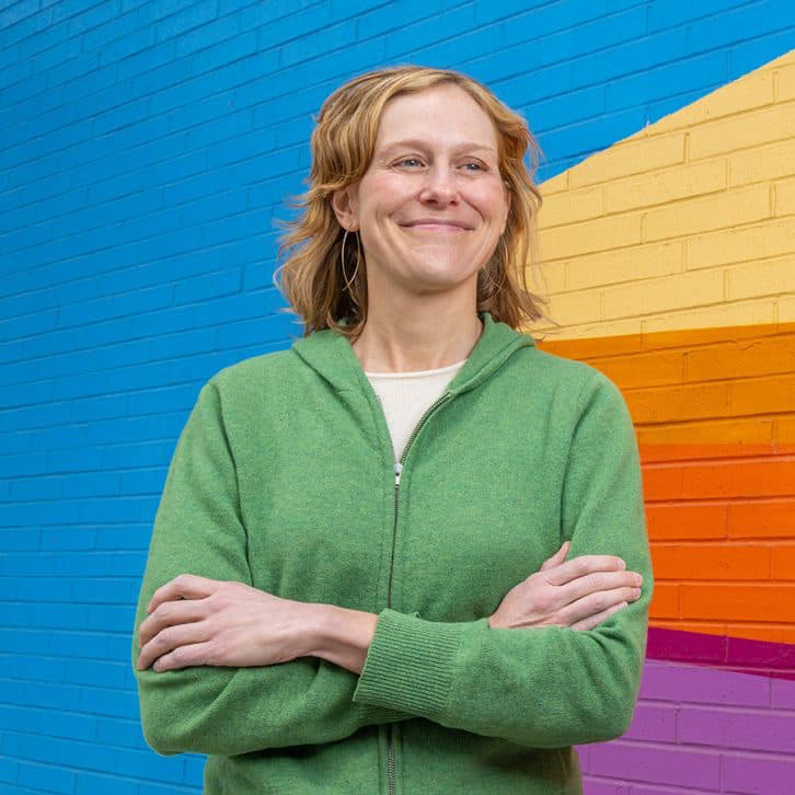 Grace Hood smiling with arms crossed in front of a brightly colored brick wall.