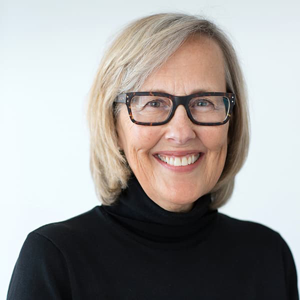 Julie snow wearing a black turtleneck sweater, dark, square rimmed glasses with shoulder-length blonde hair, and smiling with her teeth.