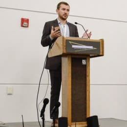 Image of Tyler Svitak in a dark suite standing at a lectern speaking.