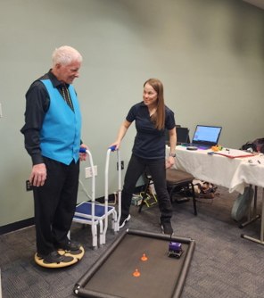 An older adult stands on a balance board which controls a small robot